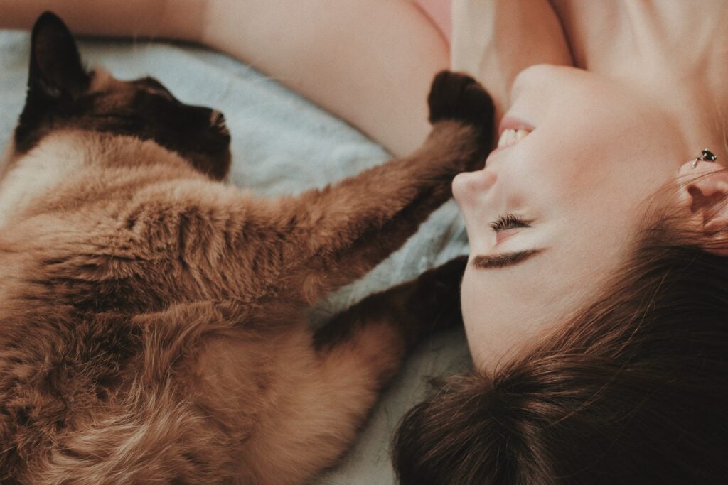 Smiling woman laying down with cat who is touching her face lovingly.